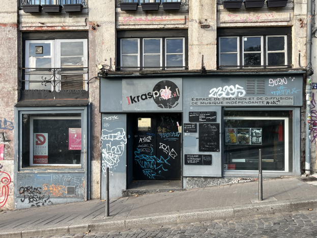 Ein mehrstöckiges Gebäude mit sichtbaren Fenstern, Geländern, Türen, Texttafeln, Plakaten, Pfählen und Graffiti an der Seite, das sich über einer Straße befindet.