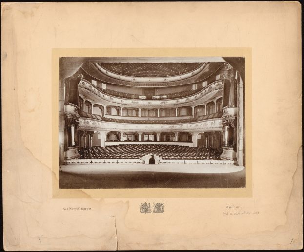 Altes Schwarz-Weiß-Foto des Auditoriums der Royal Albert Hall in London, das die Bühne, Reihen von Stühlen, Säulen, Geländer und Text mit einem Logo unten zeigt.