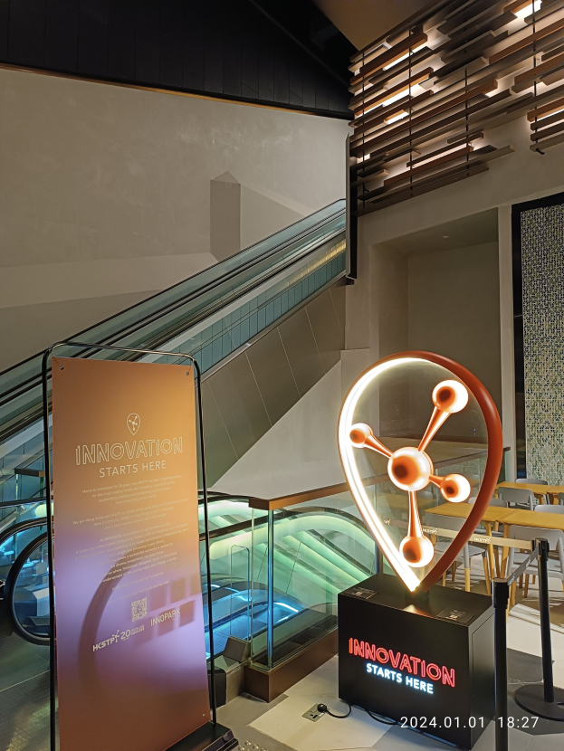Großes Schild in einem Gebäude mit einer Rolltreppe im Hintergrund, flankiert von Tischen und Stühlen auf der rechten Seite und einer Tafel mit der Aufschrift "Innovation beginnt hier" auf der linken Seite, was die Teilnahme an der HSTP20 Expo anzeigt.