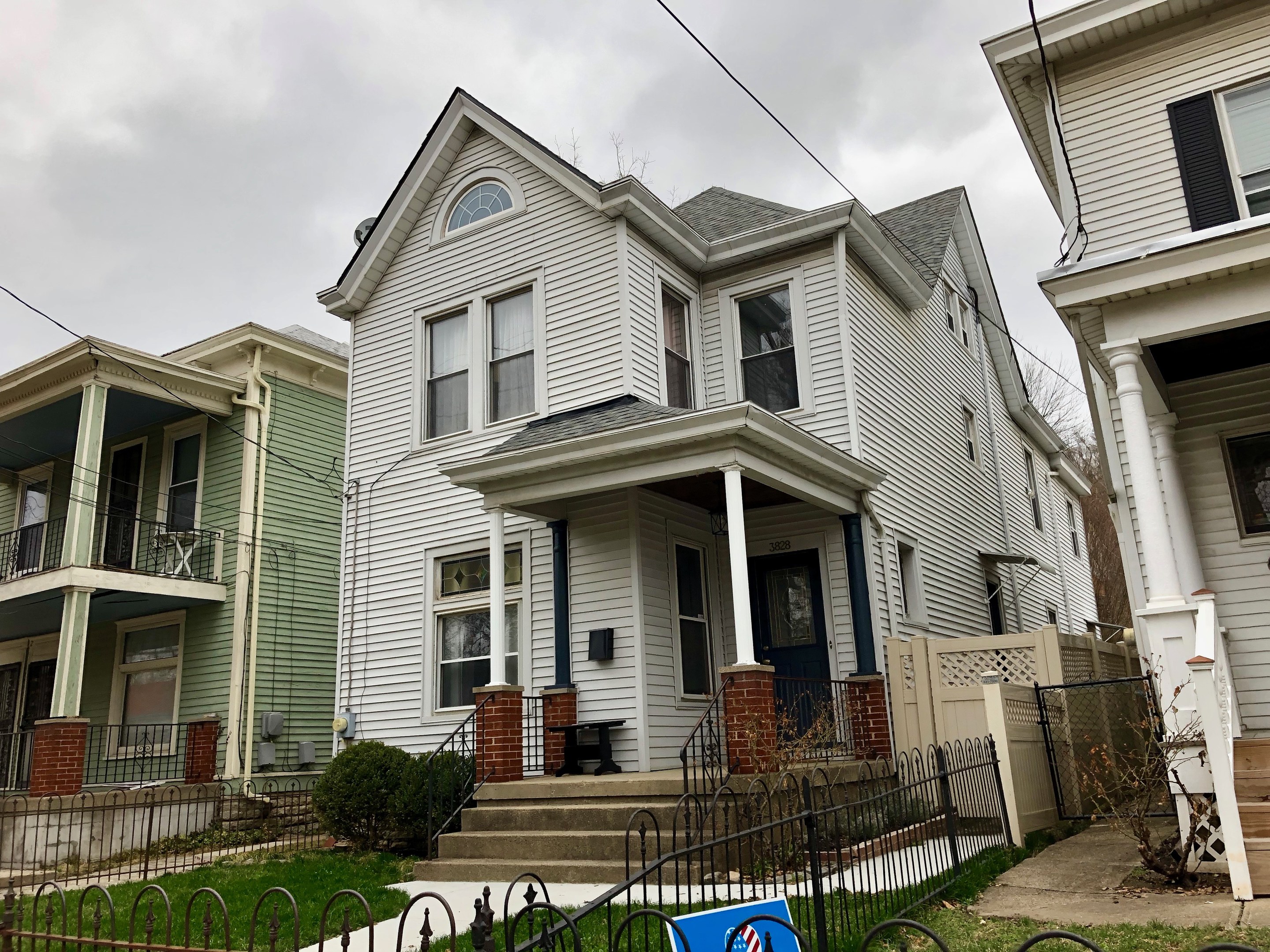 Reihe von Häusern mit Fenstern, Türen, Säulen, Geländern, Stufen und Dächern, umgeben von einem Metallzaun, Gras, Pflanzen, Bäumen und einem bewölkten Himmel, mit einem Mietschild unten, das die Adresse und die Listennummer angibt.