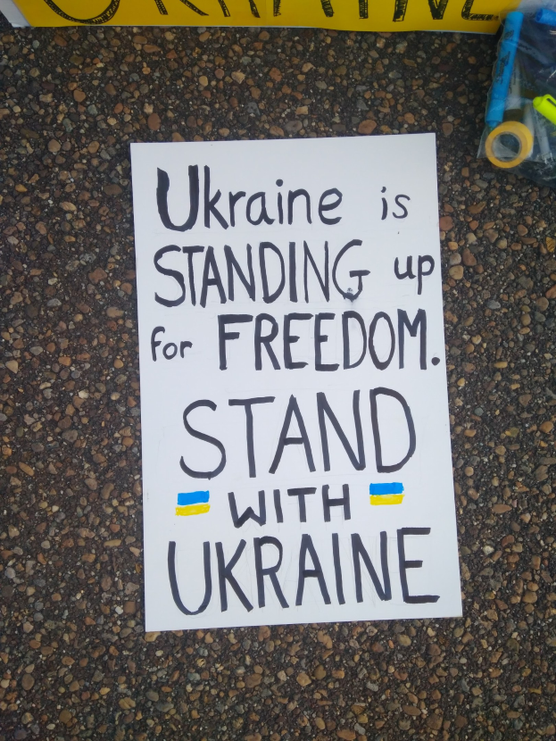 Ein weißes Blatt Papier mit dem Text "Ukraine steht für die Freiheit ein, steht mit Ukraine" auf dem Boden vor einer gelben Fahne platziert, mit einer Plastikhülle mit verschiedenen Gegenständen rechts daneben.