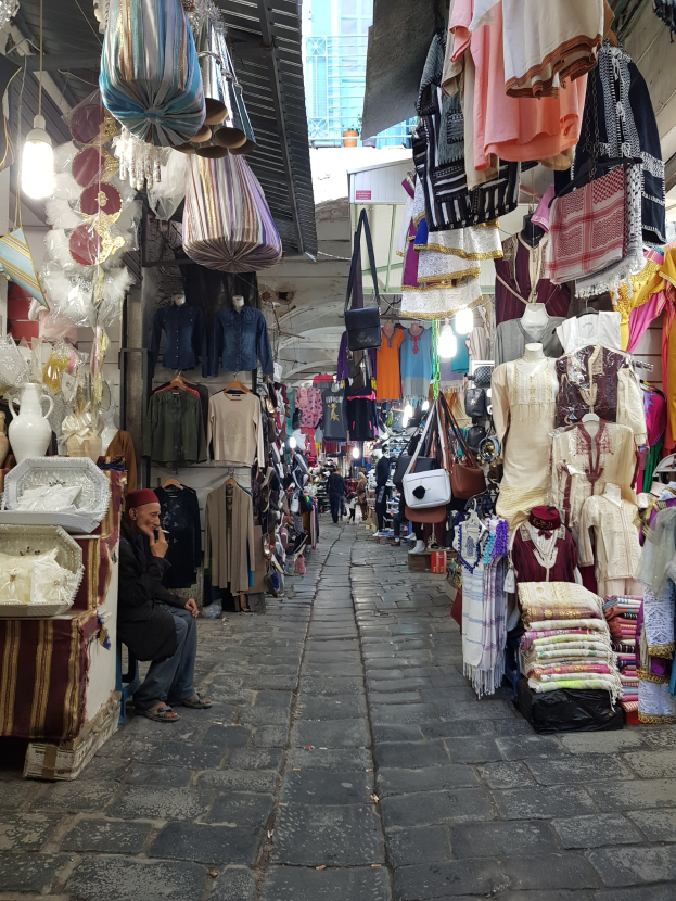 Ein belebter Straßenmarkt in der Altstadt von Jerusalem mit Ständen, die Kleidung, Taschen, Lampen und andere Artikel verkaufen, während Menschen umhergehen und die Stände durchstöbern, mit einem Gebäude im Hintergrund.