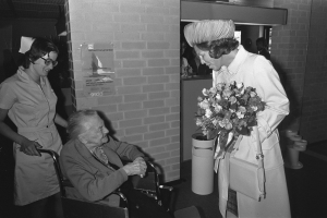 Eine ältere Frau im Rollstuhl, die einen Blumenstrauß hält, wird von einer lächelnden Krankenschwester begrüßt, mit einem Plakat, Mülltonne und ein paar anderen Menschen im Hintergrund, alles in Schwarz-Weiß.