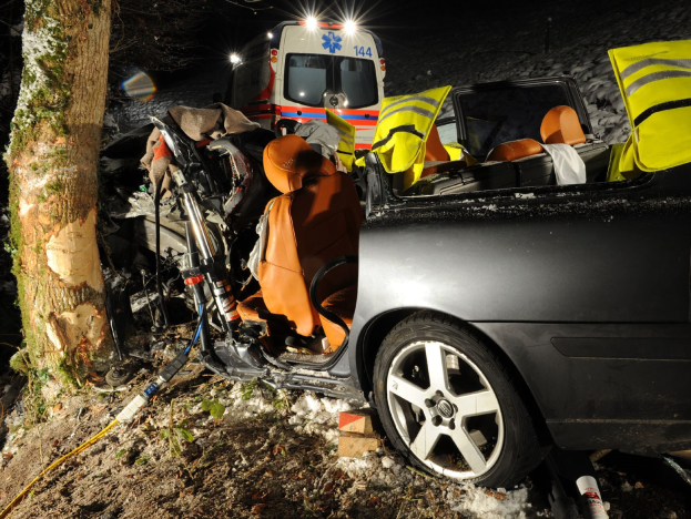 Ein beschädigtwagen am Rand einer schneebedeckten Straße mit einem Krankenwagen im Hintergrund, umgeben von Bäumen.