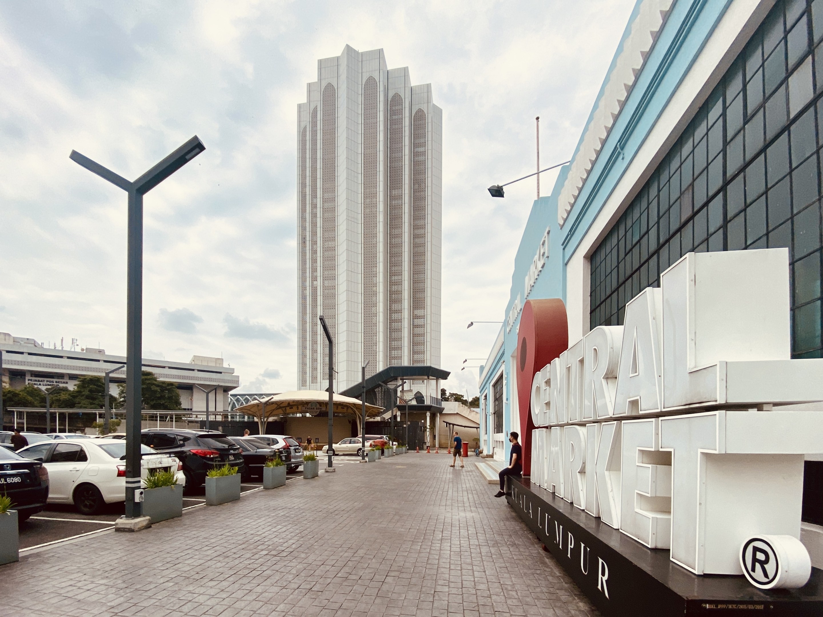 Ein belebter zentraler Markt in Kuala Lumpur, Malaysia, mit hohen Gebäuden, Fahrzeugen, Fußgängern, Straßenmöbeln und Bäumen unter einem bewölkten Himmel.