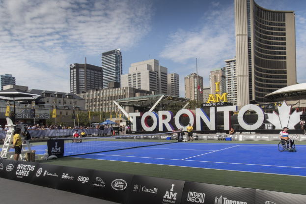 Gruppe von Menschen, die Tennis auf einem Court mit einer Stadtkulisse im Hintergrund spielen, umgeben von sitzenden und stehenden Zuschauern, Bannern, Gebäuden, Bäumen, Polen, Fahnen und einem bewölkten Himmel.