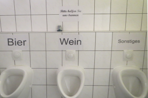 Drei weiße Toiletten an einer Wand montiert mit Schildern darüber, die jeweils "Bier Wein" und "Sonstiges" lesen.