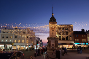 Eine belebte Stadtstraße mit Fußgängern, Fahrzeugen, Fahrrädern, Mülltonnen, Gebäuden, Laternen, Verkehrsampeln und einem Uhrenturm im Hintergrund, geschmückt mit Weihnachtslichtern am Himmel.