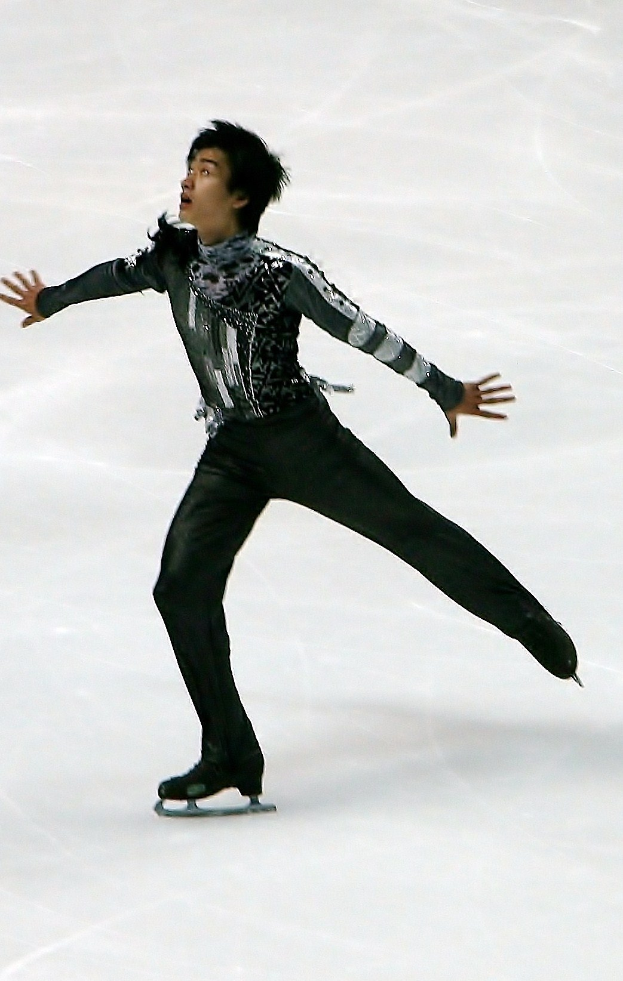 Männlicher Eiskunstläufer in einem schwarzen Kleid, der über das Eis gleitet und eine Choreographie mit synchronen Arm- und Beinbewegungen präsentiert.