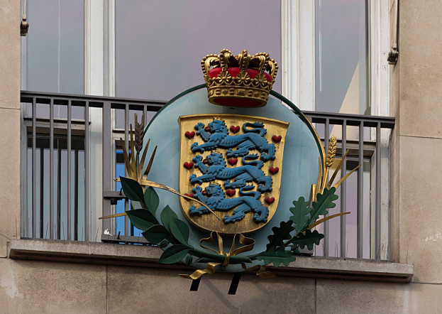 Wappen der britischen Königsfamilie auf einem Gebäude mit einer Krone und Blätter-Logo, Fenstern und Gittern im Hintergrund.