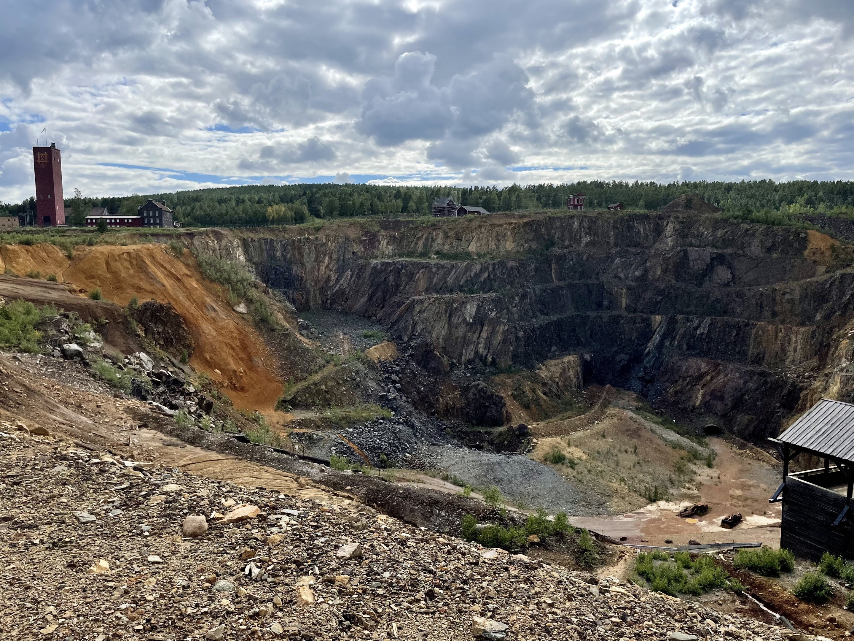 Großer Tagebau inmitten eines Schotterfeldes umgeben von Bäumen, Gebäuden und einer Hütte, mit bewölktem Himmel und einem mit Steinen und Pflanzen bedeckten Boden.