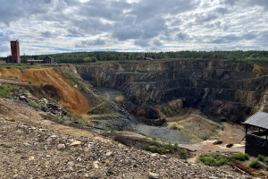 Großer Tagebau inmitten eines Schotterfeldes umgeben von Bäumen, Gebäuden und einer Hütte, mit bewölktem Himmel und einem mit Steinen und Pflanzen bedeckten Boden.