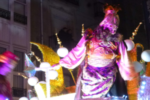 Ein Mann in einem Kostüm steht auf einem dekorierten, beleuchteten Festwagen in einer Stadtparade, mit einem Gebäude mit Fenstern und Geländern im Hintergrund.