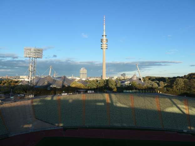 Das Olympische Stadion in Berlin, Deutschland, mit dem Fernsehturm im Hintergrund, umgeben von Bäumen, Gebäuden und Lichtern, unter einem bewölkten Himmel.