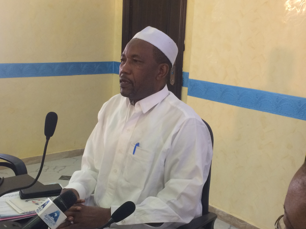 Ein Mann in einem weißen Gewand und Hut sitzt an einem Tisch mit Mikrofonen, spricht, während Papiere und Gegenstände auf dem Tisch sichtbar sind, mit einer anderen Person mit Brille zu seiner Rechten, in einem Raum mit einer Wand und einer Tür im Hintergrund.