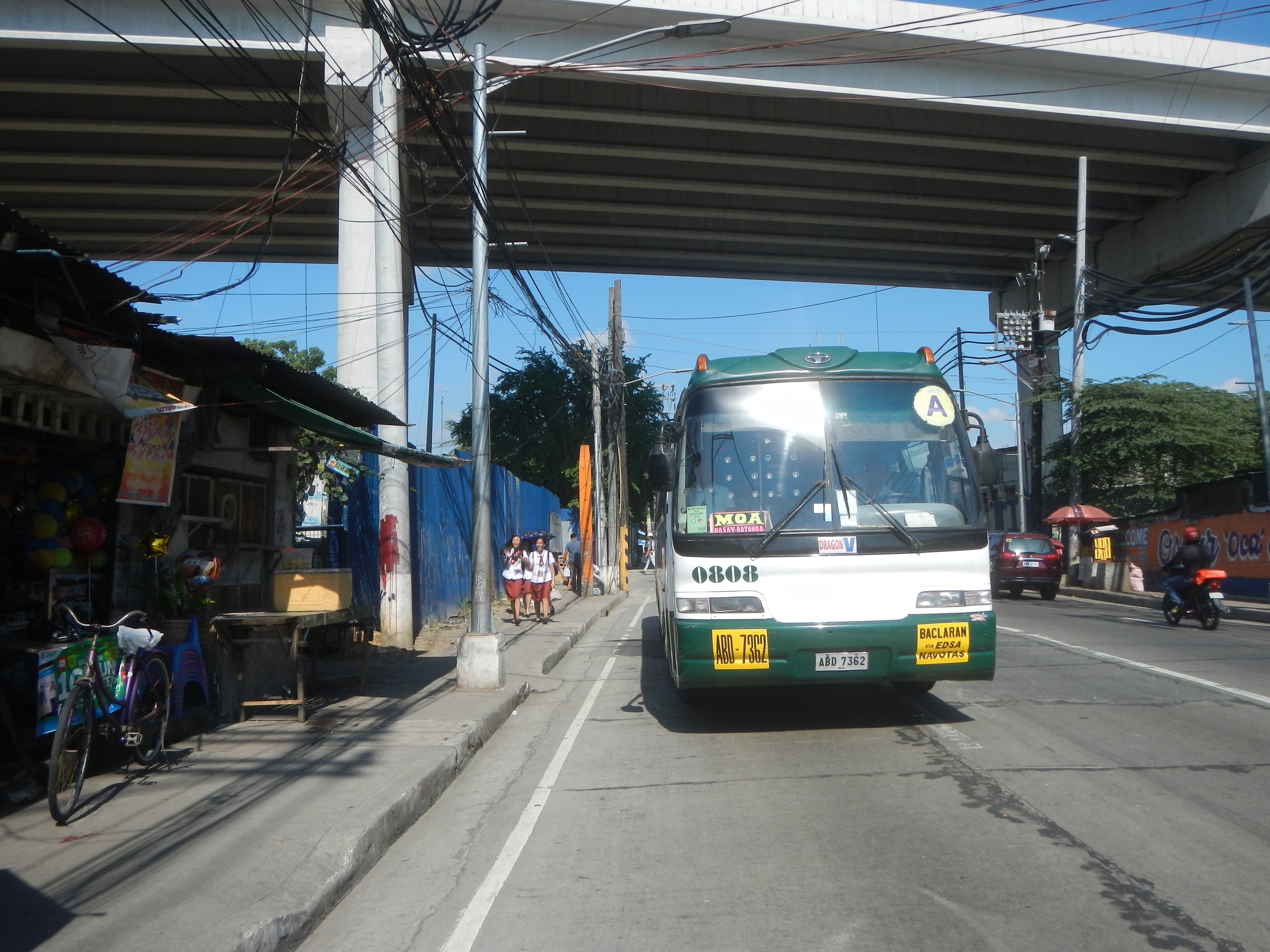 Ein grüner und weißer Bus fährt auf einer Straße neben einer Brücke, mit Strommasten und -leitungen entlang der Straße, Fußgängern auf dem Gehweg, parkenden Fahrrädern und Geschäften an der Straße, unter einem klaren blauen Himmel mit Bäumen im Hintergrund.