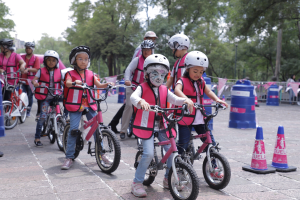 Kinder mit Helmen fahren auf einer von Verkehrskegeln markierten Straße Fahrräder, einige mit Gesichtsbemalung, vor einem Hintergrund aus Bäumen und einem klaren Himmel.