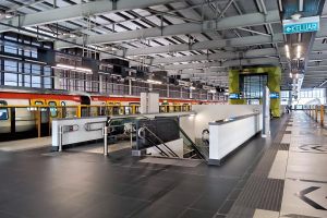 Ein Bahnhof mit einem Zug auf den Schienen, ein Bahnsteig mit Geländern, Treppen mit Handläufen, Schilder mit Text, Lampen an der Decke und Glaswände.