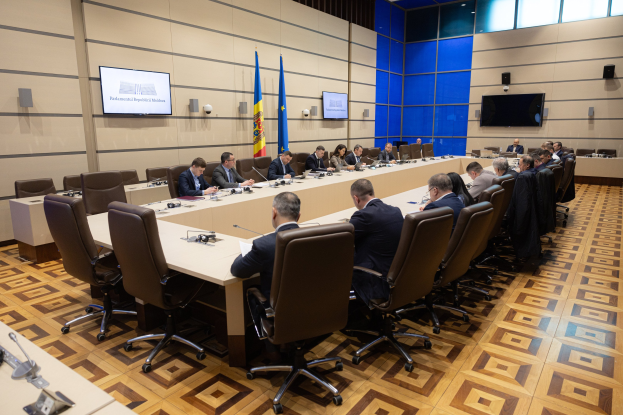 Große Gruppe von Menschen an einem langen Tisch mit Mikrofonen und Papieren in einem Raum mit Flaggen, Fernsehern, Deckenleuchten und Lautsprechern, was auf eine Sitzung des Europäischen Parlaments in Brüssel hinweist.
