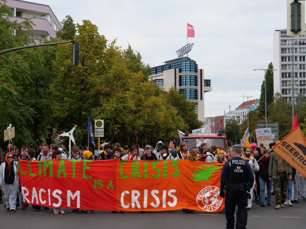 Eine Gruppe von Menschen marschiert auf einer Straße, hält ein Banner mit der Aufschrift "Klima-Krise ist eine Krise", mit Bäumen, Laternenpfählen und Schildern an der Straße, einem geparkten Fahrzeug in der Nähe und Gebäuden im Hintergrund unter einem klaren blauen Himmel.
