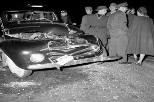 Eine Schwarz-Weiß-Szene eines beschädigten Autos am Straßenrand, umgeben von einer Gruppe besorgter Menschen.