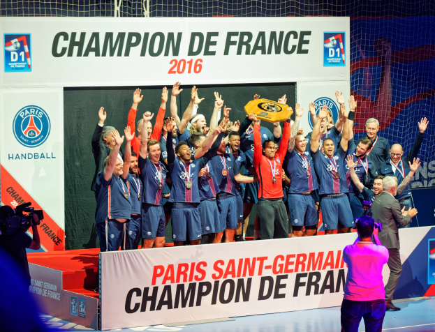 Eine Gruppe von Männern hält einen Pokal auf einer Bühne, während eine Videokamera aufnimmt und ein "Champion de France 2016"-Banner im Hintergrund zu sehen ist.