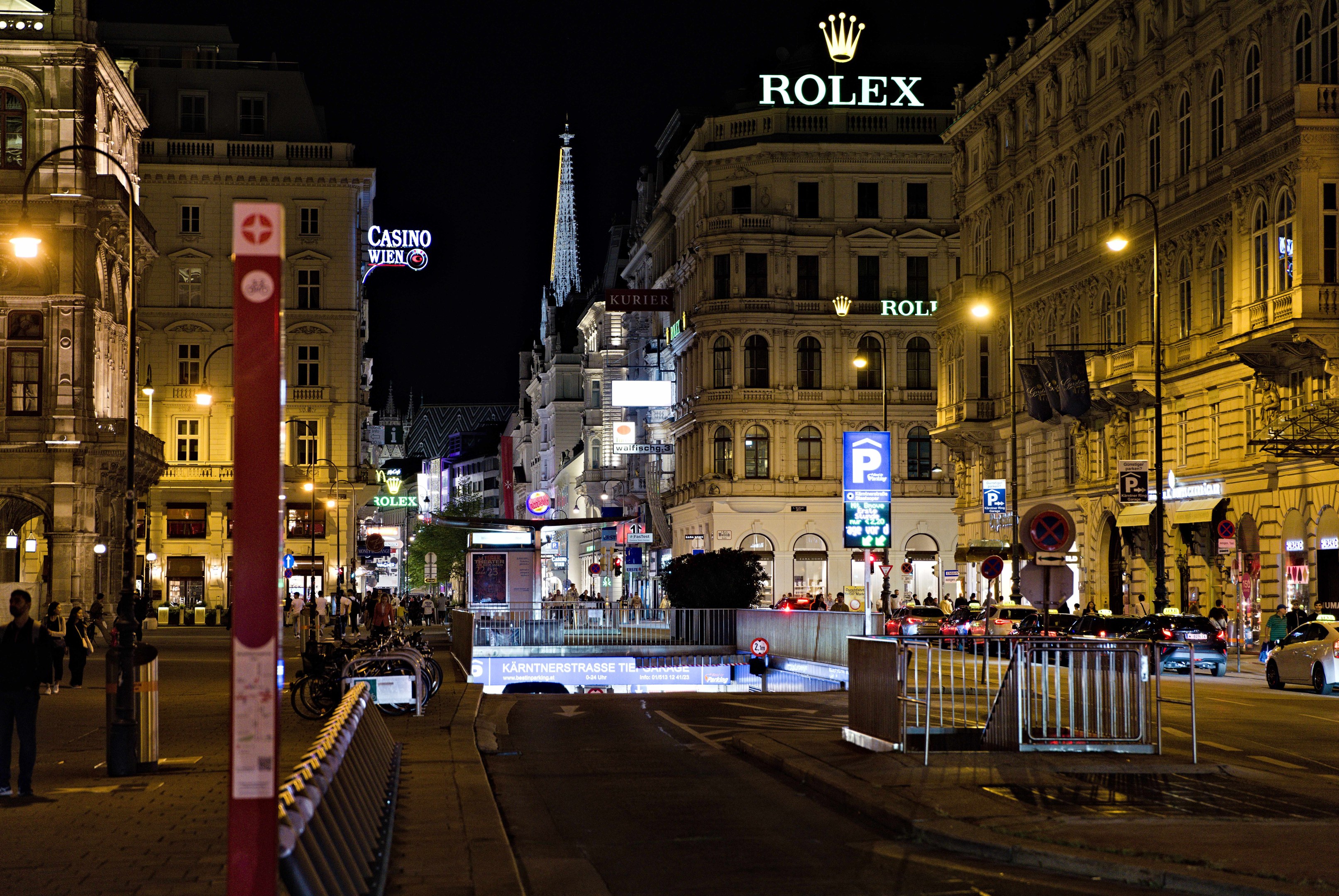 Eine nächtliche Stadtstraße mit fahrenden Autos, Fußgängern auf den Gehwegen, parkenden Fahrrädern, Straßenlaternen, hohen Gebäuden, Absperrungen, Polen und einem Rolex-Logo auf einem Gebäude im Hintergrund.