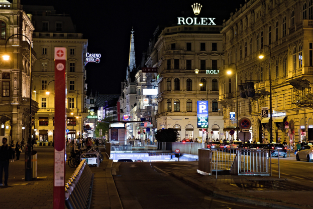 Eine nächtliche Stadtstraße mit fahrenden Autos, Fußgängern auf den Gehwegen, parkenden Fahrrädern, Straßenlaternen, hohen Gebäuden, Absperrungen, Polen und einem Rolex-Logo auf einem Gebäude im Hintergrund.
