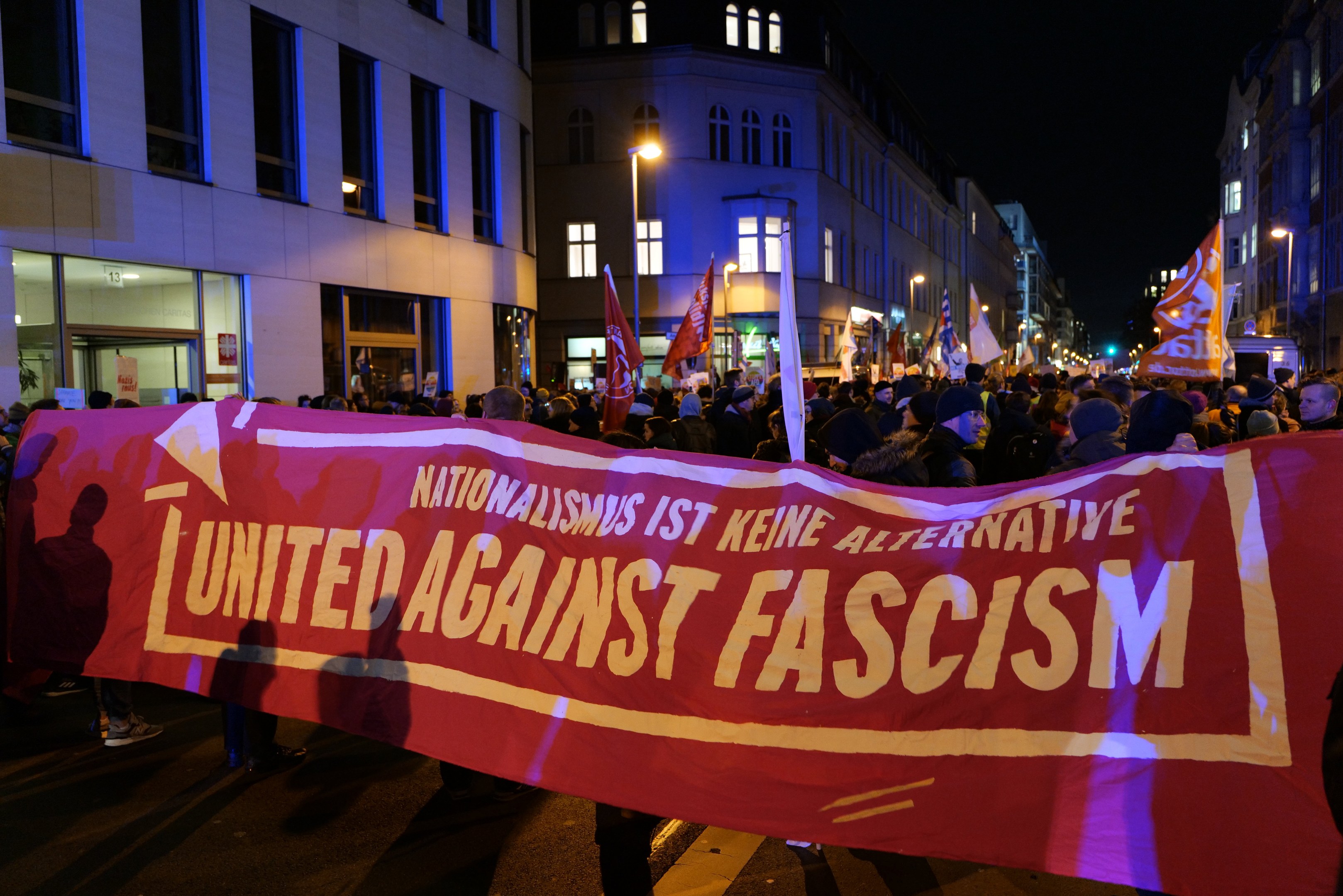 Eine Gruppe von Menschen marschiert auf einer Straße und hält ein Banner mit der Aufschrift "Vereinigt gegen Faschismus", mit Gebäuden, Laternenpfählen und Schildern an der Straße und einem sichtbaren Himmel.