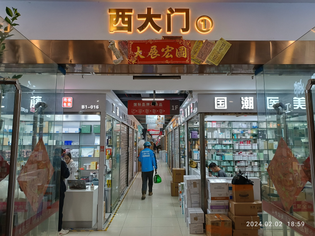Innenraum einer chinesischen Apotheke mit Glaswänden, Regalen, verstreuten Kartons und einer Pflanze auf der linken Seite; ein Schild oben zeigt an, dass es sich um eine chinesische Drogerie handelt.