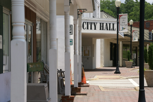 Rathaus mit Gebäuden, Straßenlaternen, Straßenschildern, Motorfahrzeugen, Absperrgittern, Stühlen, Topfpflanzen, Bäumen und einem bewölkten Himmel im Hintergrund.