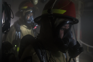 Zwei Feuerwehrleute in Helmen und Gasmasken stehen in einem dunklen Raum, mit einem Mast mit Drähten auf der linken Seite und verschiedenen Gegenständen im Hintergrund.