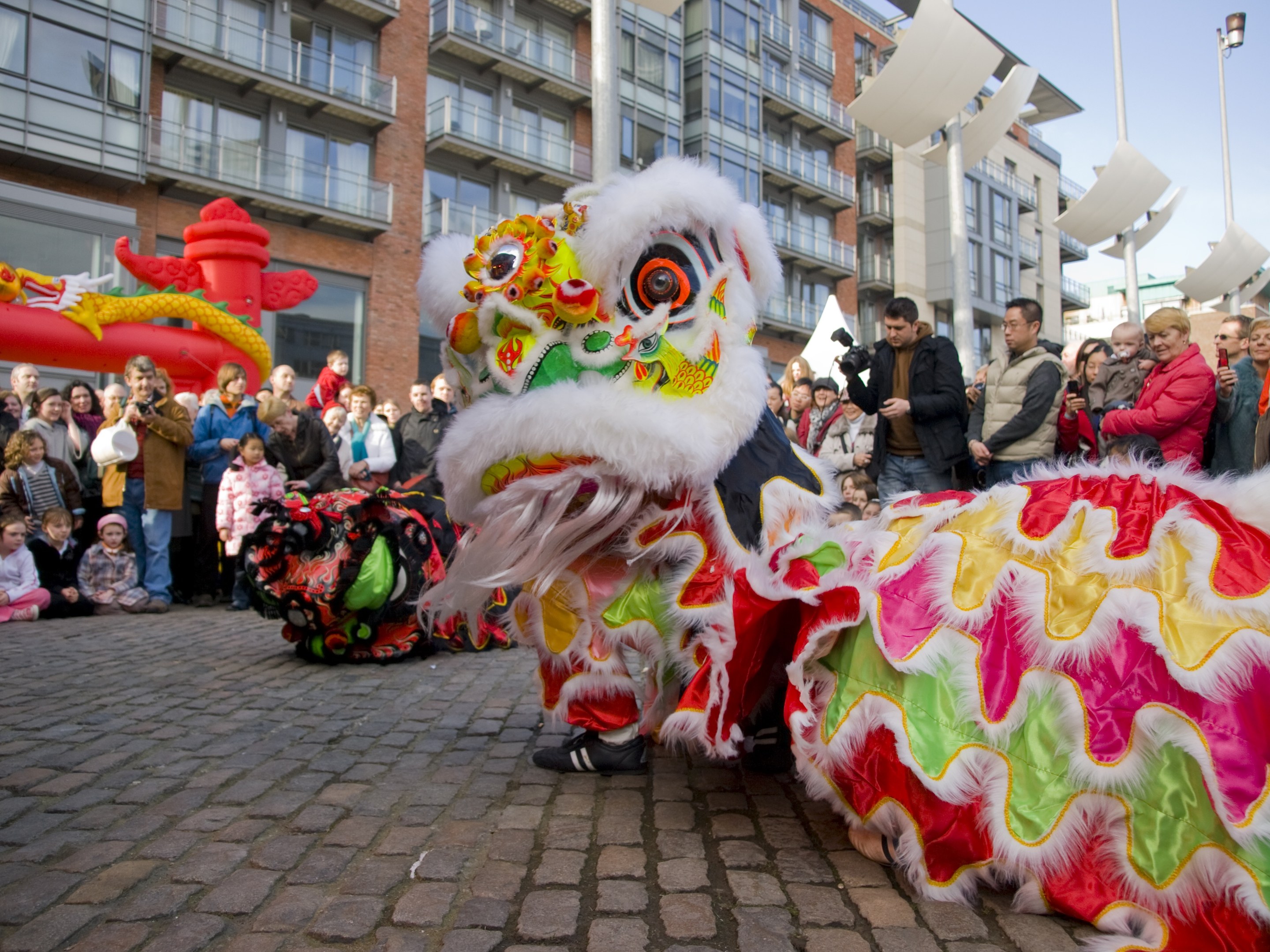 Ein lebendiges chinesisches Neujahrsfest in Amsterdam mit einer Löwen-Tanzvorstellung vor einer Zuschauermenge, einige halten Kameras, vor einem Hintergrund aus Gebäuden, Laternenmasten und einem klaren blauen Himmel.