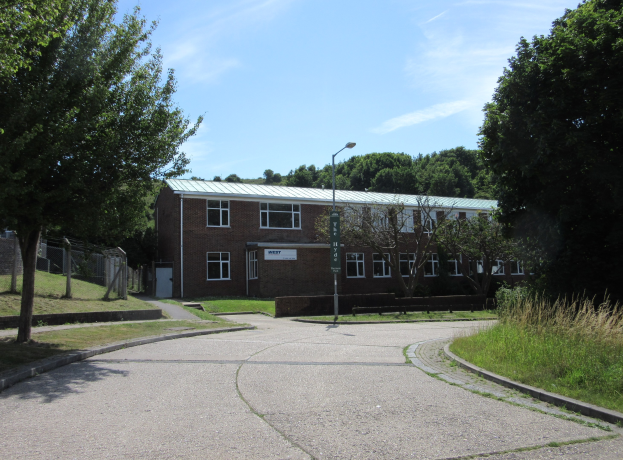 Ein Schulgebäude mit Fenstern, einem Straßenschild, einem Schild, einer Baumgruppe, Gras, einem Zaun und einem bewölkten Himmel, das sich in einem Park mit saftigem Gras befindet.
