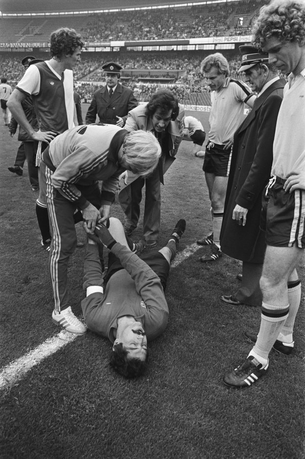 Schwarzes und weißes Bild eines Fußballspielers auf dem Boden umgeben von einer Gruppe von Männern, mit einer Menschenmenge im Hintergrund eines Stadions.