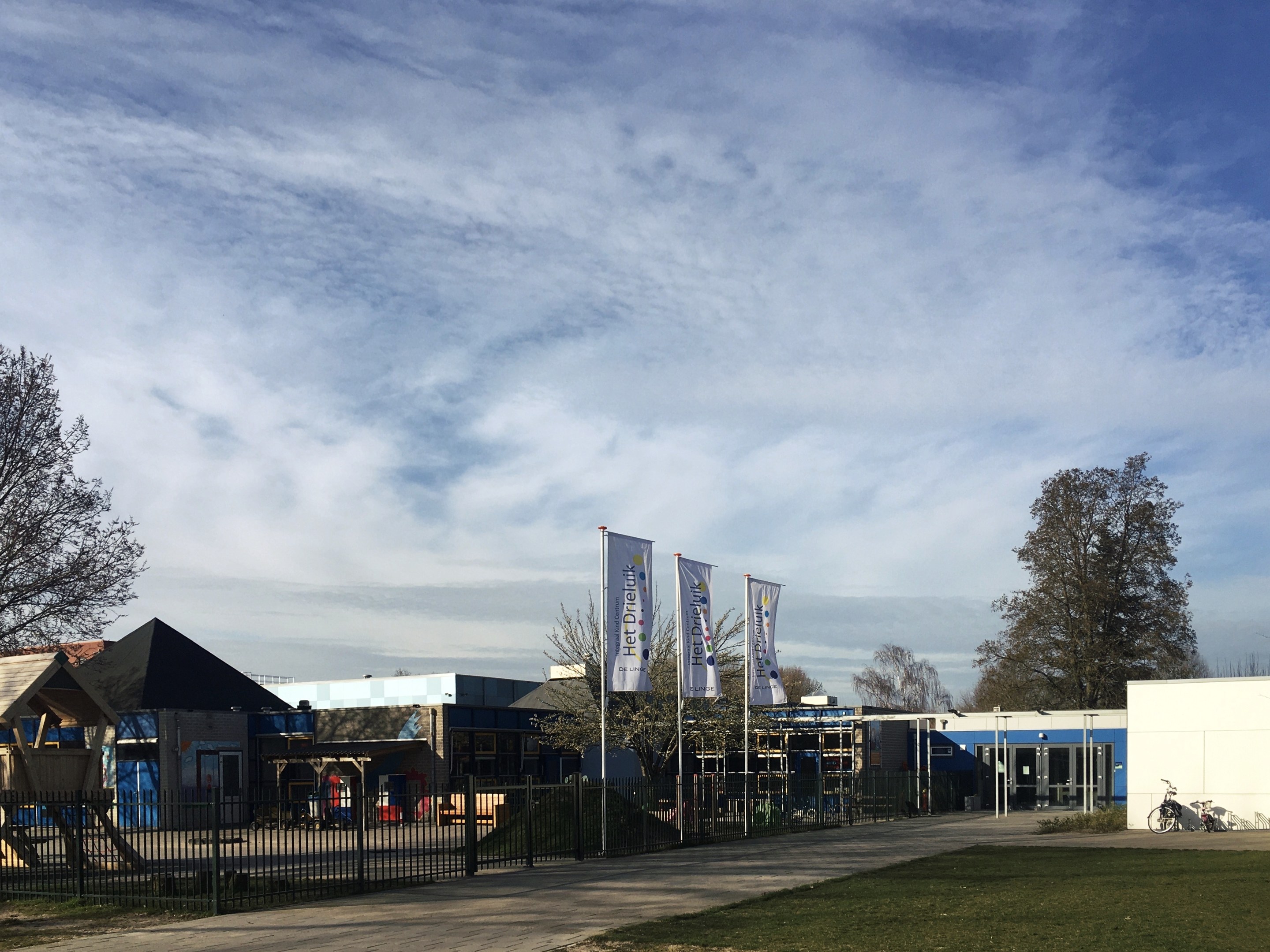 Großes Gebäude mit Metallzaun, Weg, Bäumen, Bannern, einem geparkten Fahrrad, nahen Häusern und einem bewölkten Himmel, identifiziert als der neue Spielplatz an der Schule.