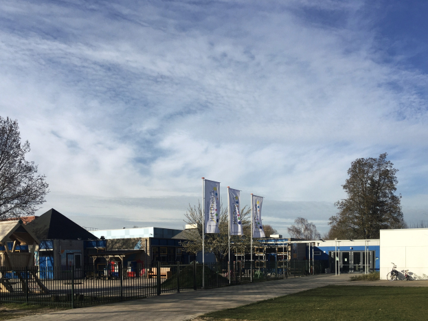 Großes Gebäude mit Metallzaun, Weg, Bäumen, Bannern, einem geparkten Fahrrad, nahen Häusern und einem bewölkten Himmel, identifiziert als der neue Spielplatz an der Schule.