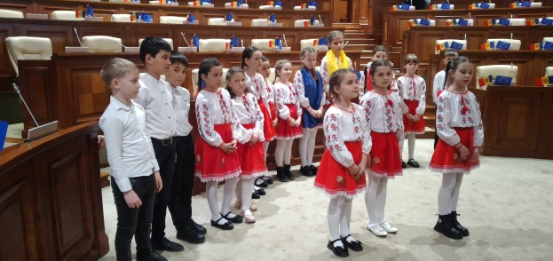 Eine Gruppe von Kindern in weißen und roten Kleidern steht zusammen in einem Raum mit einem Podium und einem Mikrofon links, Stühlen und Tischen im Hintergrund und Flaggen, die eine nationale Versammlung anzeigen.