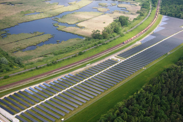 Luftaufnahme einer Solaranlage mit Solarpanelen auf einem Feld, umgeben von Bäumen, Gras, Pflanzen und Wasser, mit einem Zug auf einer nahen Bahnlinie.