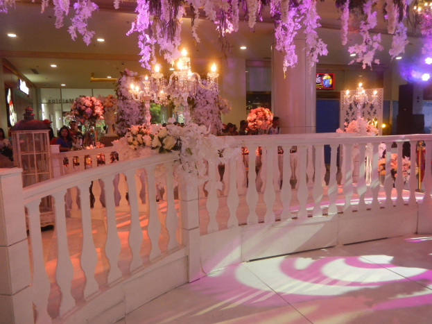 Weiße Reling mit Blumen und Kronleuchtern hängt von der Decke bei einer Hochzeitsfeier, mit Menschen drumherum, Säulen im Hintergrund, beleuchtete Lichter und ein Bildschirm auf der rechten Seite.