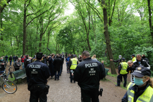 Eine Gruppe von Polizisten steht vor einer Menschenmenge, einige tragen Mützen und Masken, mit Fahrrädern und einer Bank im Vordergrund und Bäumen und Himmel im Hintergrund, während einer Anti-Terror-Demonstration in Berlin.