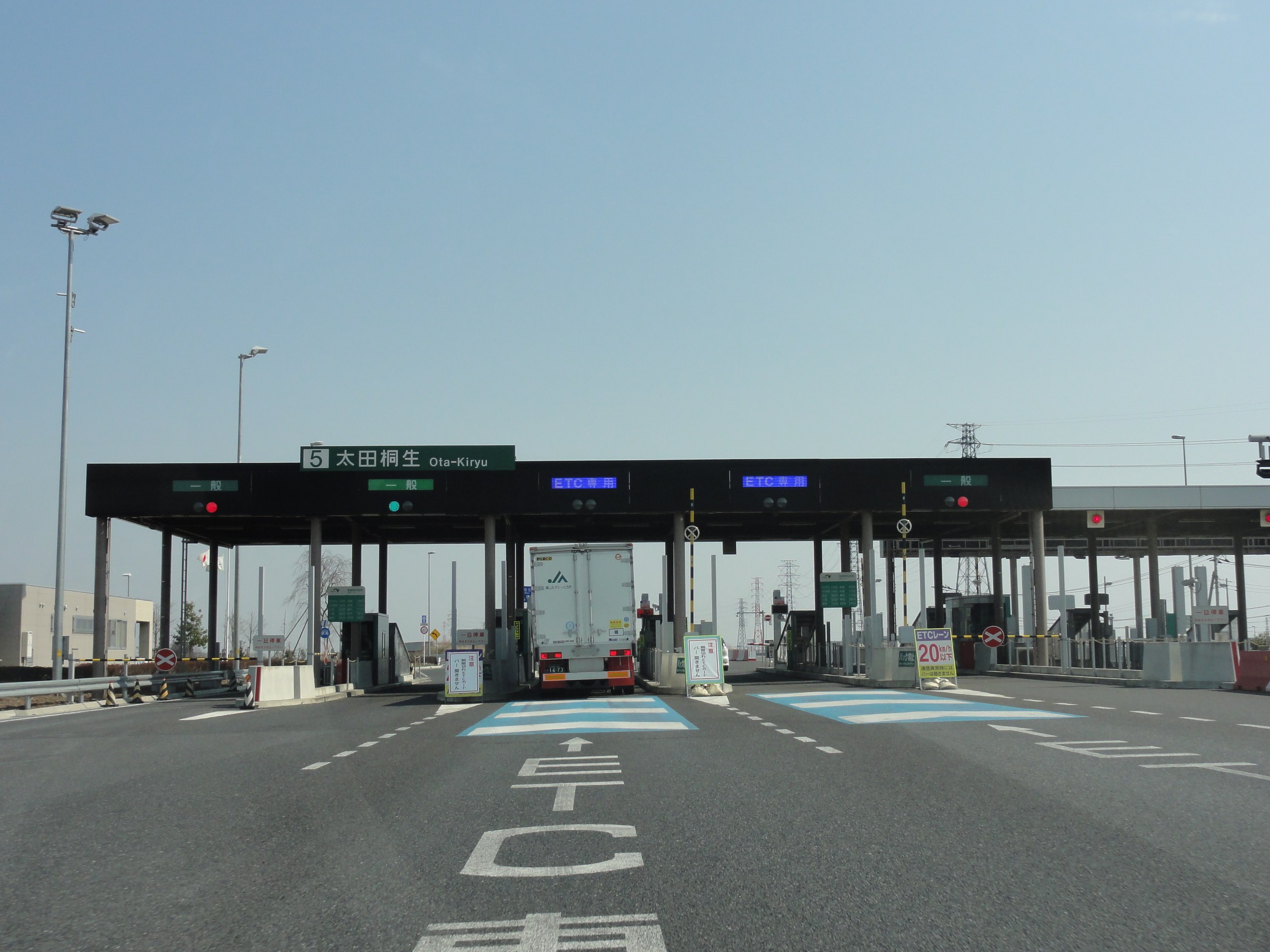 Mautstation an der Straße mit einem vor ihr geparkten Lastwagen, weiße und blaue Straßenmarkierungen, Laternenpfähle, Schilder, Gebäude, Bäume und einen klaren blauen Himmel im Hintergrund.