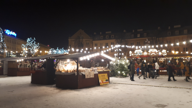 Ein verschneiter nächtlicher Weihnachtsmarkt mit Menschen, beleuchteten Buden, Pflanzen, Bäumen, Gebäuden und Schildern unter einem bewölkten Himmel.