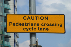 Gelbes Warnschild an einem Pfahl vor einem Gebäude, auf dem steht "Fußgänger überqueren Fahrradspur" vor einem klaren blauen Himmel.