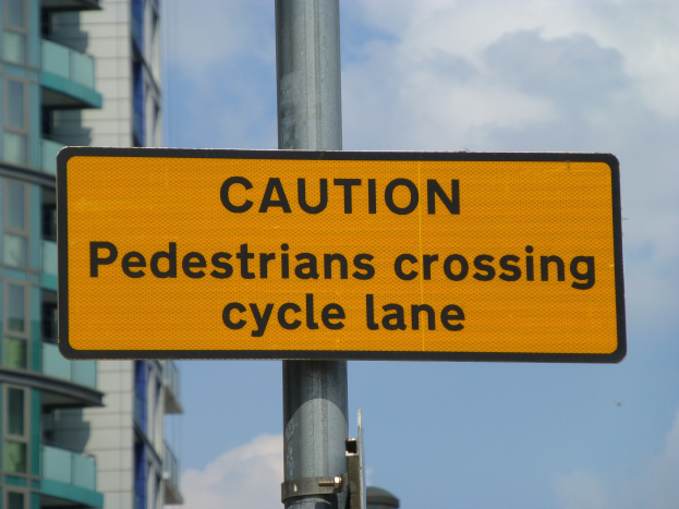 Gelbes Warnschild an einem Pfahl vor einem Gebäude, auf dem steht "Fußgänger überqueren Fahrradspur" vor einem klaren blauen Himmel.
