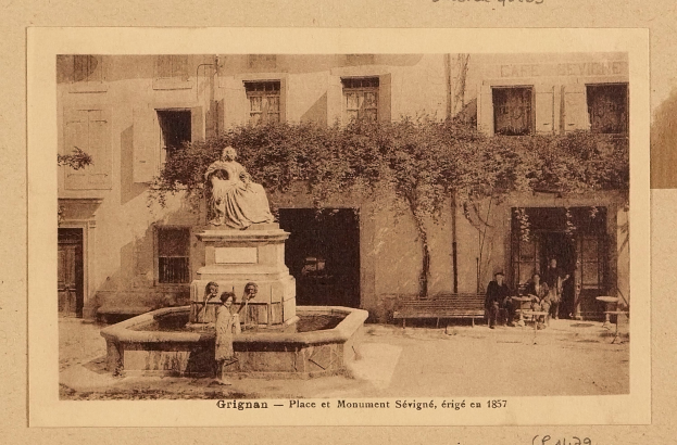 Ein altes Foto eines Brunnens vor einem Gebäude, mit einer Statue auf einem Sockel in der Mitte, Menschen auf Bänken um den Brunnen herum und einem Baum im Hintergrund, mit Text am unteren Rand des Bildes.