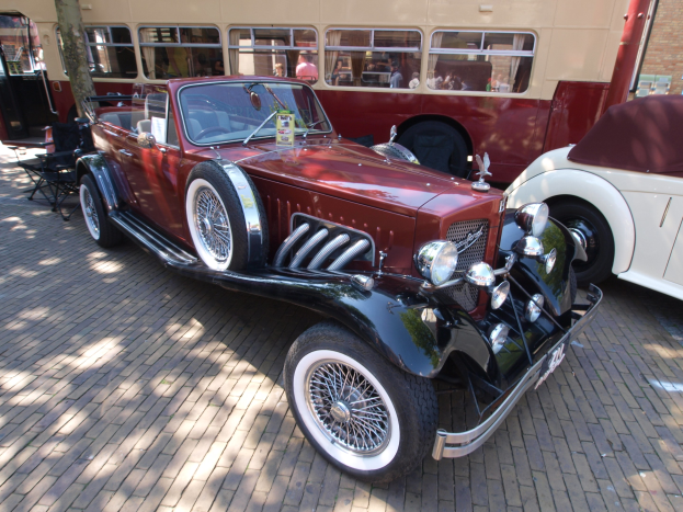 Ein Vintage-Auto neben einem Doppeldeckerbus auf einer Kopfsteinpflasterstraße geparkt, mit Stühlen und einem Baum im Vordergrund, einem Gebäude im Hintergrund und Menschen im Bus zu sehen.