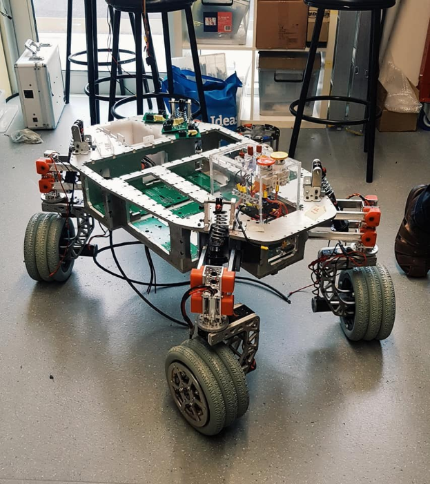 Ein Roboter sitzt auf einem Tisch in einem unordentlichen Raum mit Hockern, Kartons und anderen Gegenständen, während ein Person Bein rechts zu sehen ist.