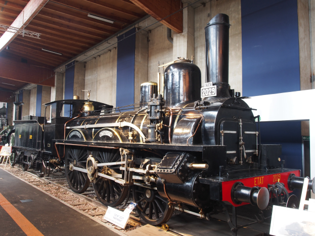 Alte Dampflokomotive in Museums Setting mit umgebenen Eisenbahnschienen, Informationsschildern und anderen Ausstellungsstücken gegen eine Wand mit Deckenbeleuchtung.