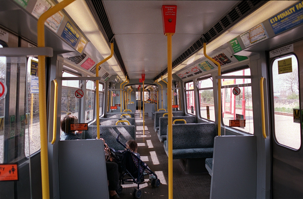 Innensicht eines öffentlichen Verkehrsmittels mit Sitzreihen, einem Kinderwagen mit Baby, offenen Fenstern und sichtbaren Hinweisschildern.